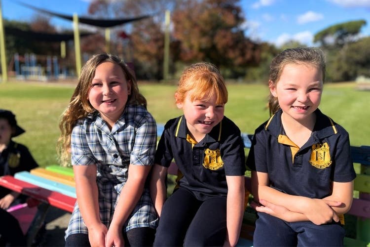 3 students, smiling and squinting in the sun as they sit on a chair in the playground.