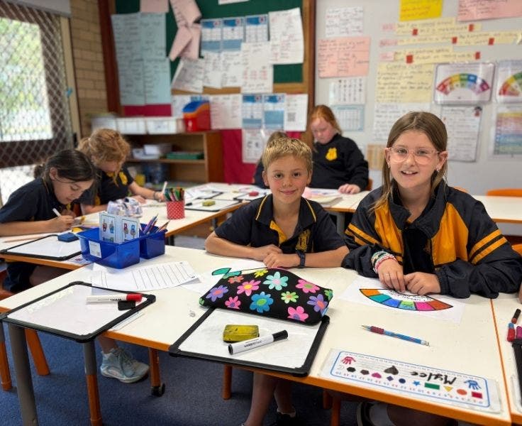 2 students sitting at their desk in class, smiling at the camera.