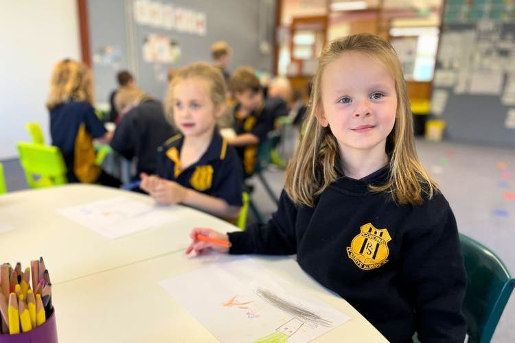 2 students sitting at their desk in class, the one in the foreground is smiling at the camera.