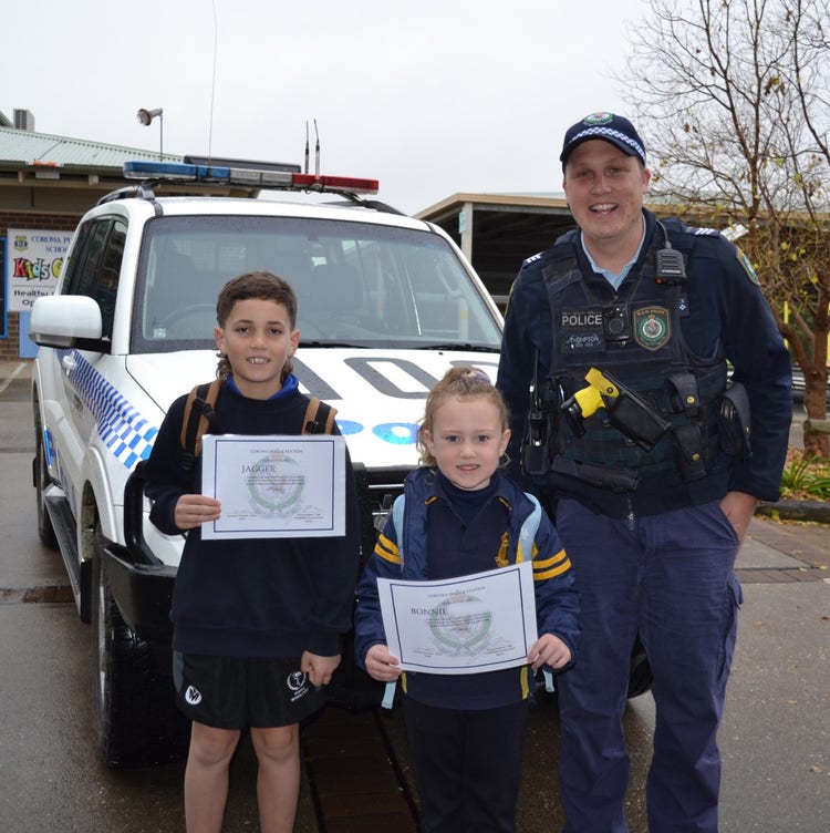 2 students holding their certificates up infront of a police car next to our 000 Program police officer.