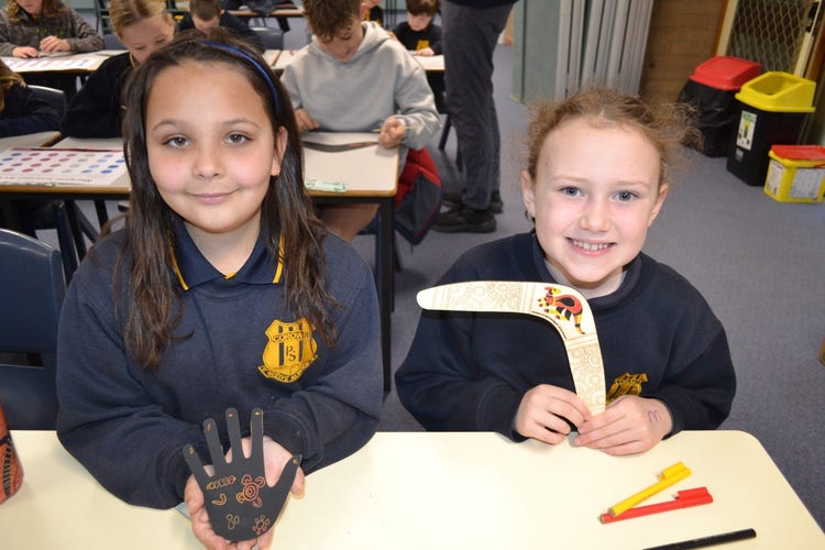 2 students smiling at the camera showing their craft activities, one is a hand artwork and the other a bangerang.