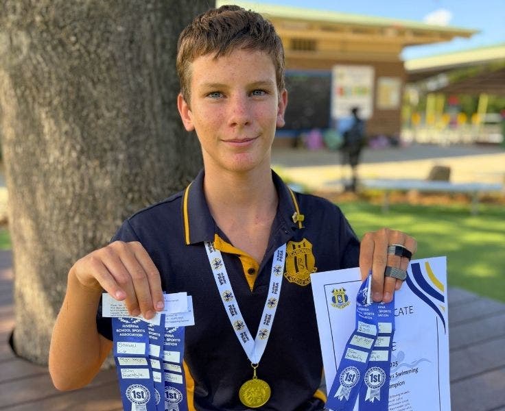 A student holding a sporting sertificate, many blue ribbons and a medal around his neck for success in swimming.