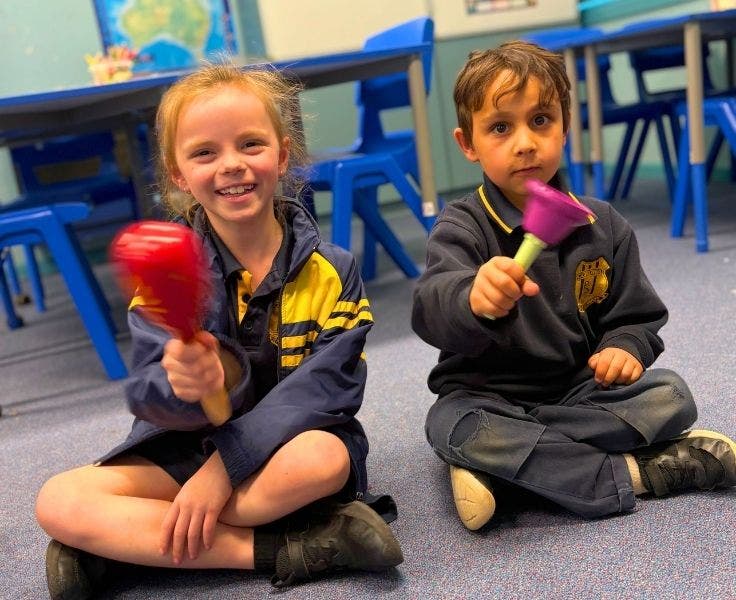 2 students sitting on the floor in the classroom shaking a maraca and a bell.