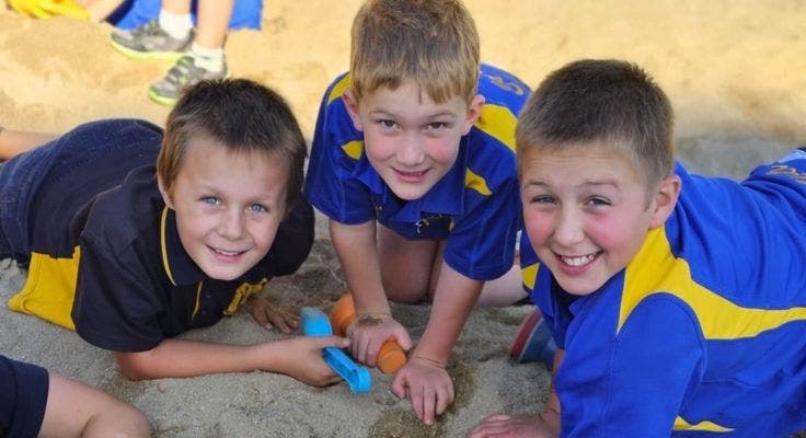 3 students digging in the sandpit, looking at the camera smiling.