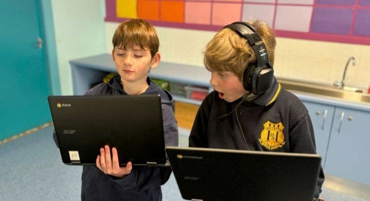 two students holding their laptops, the student on the right is wearing headphones and looking at the other students laptop, surprised.