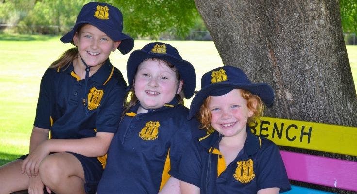 3 students sitting on a Buddy Bench in the playground, smiling at the camera.